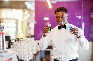 African american bartender at bar holding two coctail shots. Alcoholic beverage preparation. photo
