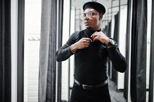 Stylish casual african american man at black outfit and beret with waist bag at fitting room clothes store , looking at mirror. photo