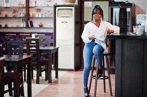 Stylish african american women in white blouse and blue jeans posed at cafe with newspaper. photo