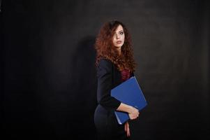 Brunette curly haired girl in black office jacket with skirt, on bra with folder of documentts at hands on studio against black background. Sexy businesswoman. photo