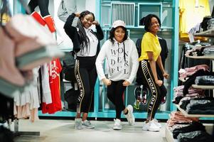 Three afican american women in tracksuits shopping at sportswear mall against shelves. Sport store theme. photo