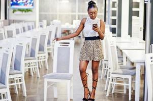 encantadora mujer afroamericana con falda de leopardo posando en el restaurante con teléfono móvil. foto