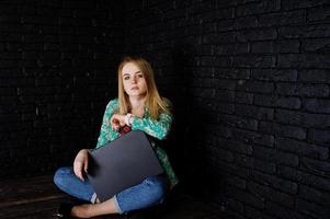 Stylish blonde girl in jacket and jeans with laptop against brick black wall at studio. photo