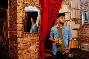 Portrait of handsome successful bearded south asian, young indian freelancer in blue jeans shirt standing in cafe with cocktail against mirror. photo