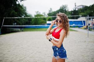 Sexy curly model girl in red top, jeans denim shorts, cup and sunglasses posed at beach volleyball field. photo