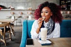 Attractive african american curly girl sitting at cafe with latte and eating cookie. photo