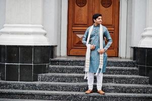 hombre indio vestido con ropa tradicional con bufanda blanca posada al aire libre contra la puerta del edificio con teléfono móvil a mano. foto