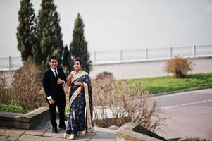 Elegant and fashionable indian friends couple of woman in saree and man in suit posed on stairs against lake. photo