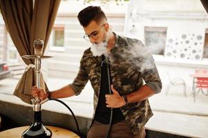 hombre árabe de barba elegante con gafas y chaqueta militar fumando narguile en el bar de la calle. modelo árabe descansando. foto