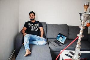 Casual beard indian man sitting at room against hookah with laptop. photo