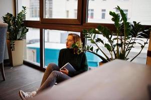alegre joven hermosa mujer pelirroja con gafas, suéter verde de lana caliente, usando su cuaderno, mientras se sienta en el café contra la ventana. se sienta en el suelo y piensa en algo. foto