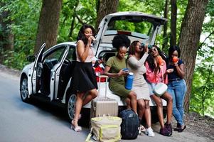 Group of five happy african american traveler girls sitting in car open trunk and drink tea from thermos. photo