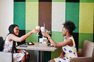 Two black african girlfriends at summer dresses drinking and cheering milkshake cocktails at cafe. photo