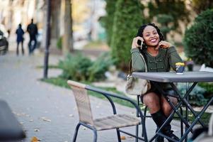 Stylish fashionable african american women in green sweater and black skirt posed outdoor cafe, sitting by table with cup of coffee and speak on mobile phone. photo