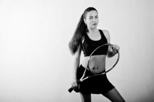 Black and white portrait of beautiful young woman player in sports clothes holding tennis racket while standing against white background. photo