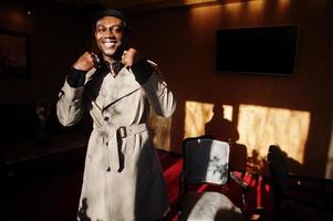 Handsome african american man posing  inside room with sunlight shadows in black hat and beige coat. photo