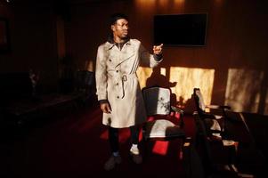 Handsome african american man posing  inside room with sunlight shadows in black hat and beige coat. photo