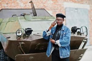 African american man in jeans jacket, beret and eyeglasses, speaking on phone against btr military armored vehicle. photo