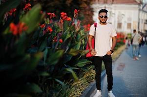 Stylish indian beard man at pink t-shirt, sunglasses and backpack. India model posed outdoor at streets of sunset city. photo