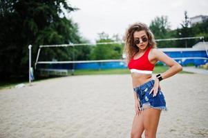 chica sexy modelo rizada en top rojo, pantalones cortos de mezclilla, copa y gafas de sol posadas en el campo de voleibol de playa. foto