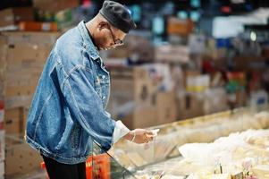 Stylish casual african american man at jeans jacket and black beret holding basket, standing near cheese fridge and shopping at supermarket. photo