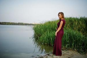 Blonde sensual woman in red marsala dress posing against lake with reeds. photo