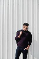 Fashion rich beard Arab man wear on turtle neck and sunglasses posed against green and white steel wall with jacket on hand. Stylish, succesful and fashionable arabian model guy. photo
