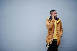 hombre africano con chaqueta en clima frío posado al aire libre contra una pared gris, hablando por teléfono móvil. foto