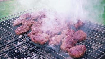 Barbeque footage shows a beautiful day in the garden where a barbecue grill is set up. Meat slices fill the grill and smoke slowly comes up from the grill and spreads. Use this to establish an outdoor video