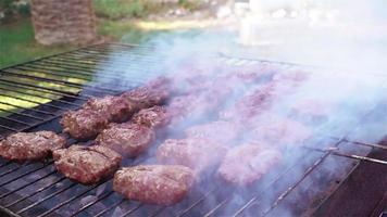 Barbeque footage shows a beautiful day in the garden where a barbecue grill is set up. Meat slices fill the grill and smoke slowly comes up from the grill and spreads. Use this to establish an outdoor video