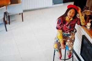 Enthusiastic african american woman in trendy coloured outfit with red beret chilling in cozy cafe, sitting on bar counter. photo