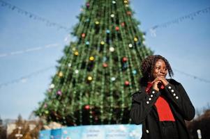 Portrait of a curly haired african woman wearing fashionable black coat and red turtleneck posing outdoor against main new year tree of city. photo