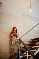 Enthusiastic african american woman in trendy coloured outfit with red beret chilling in cozy cafe, standing on stairs with cup of hot drink in hands. photo