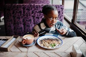 African woman in checkered cape and eyeglasses sitting at cafe and eating dessert. photo