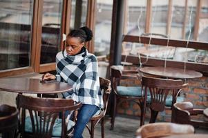 mujer africana en capa a cuadros posó en la terraza de un café al aire libre con teléfono móvil. foto