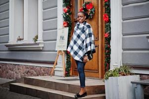 mujer africana en capa a cuadros posó al aire libre contra las decoraciones navideñas. foto