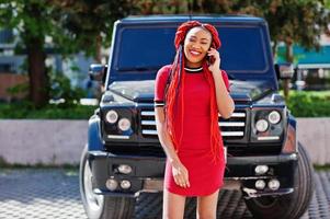 Cute and slim african american girl in red dress with dreadlocks background black mafia suv car and speking on mobile phone. Stylish black model. photo