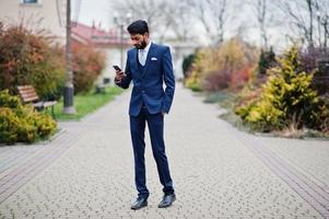 hombre de negocios indio de barba elegante con bindi en la frente, vestido con traje azul posado al aire libre mirando el teléfono móvil. foto