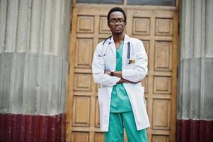 Stylish african american doctor with stethoscope and lab coat posed against door of hospital. photo