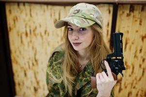 Military girl in camouflage uniform with revolver gun at hand against army background on shooting range. photo