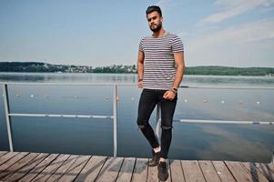 Handsome tall arabian beard man model at stripped shirt posed outdoor on pier of lake. Fashionable arab guy. photo