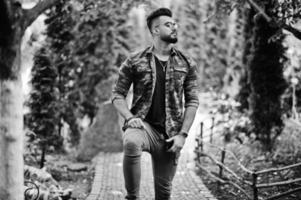 Impresionante hermoso hombre macho alto de barba árabe con gafas y chaqueta militar posado al aire libre. foto