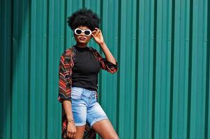 mujer africana con cabello afro, en pantalones cortos de jeans y gafas de sol blancas posadas contra una pared de acero verde. foto