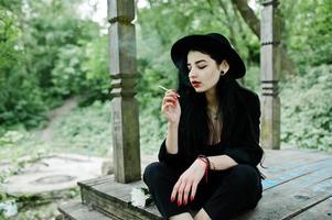 chica fumadora sensual todo en negro, labios rojos y sombrero. mujer dramática gótica fumando un cigarrillo delgado. foto