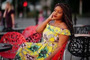 Cute small height african american girl with dreadlocks, wear at coloured yellow dress, sitting at outdoor cafe on red chair. photo