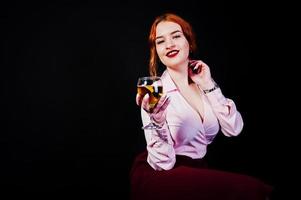 Gorgeous red haired girl in pink blouse and red skirt with glass of wine at hand isolated on black. photo