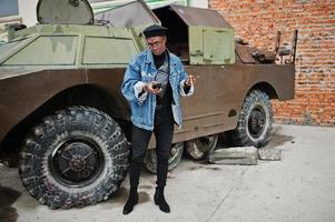 African american man in jeans jacket, beret and eyeglasses, smoking cigar and posed against btr military armored vehicle, with mobile phone at hand. photo