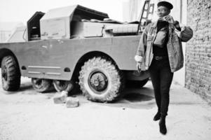 African american man in jeans jacket, beret and eyeglasses, smoking cigar and posed against btr military armored vehicle. photo