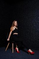 Handsome brunette girl wear on black and red high heels, sitting and posing on chair at studio against dark brick wall. Studio model portrait. photo