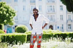 Stylish african american man in white shirt and colored pants with hat and glasses posed outdoor. Black fashionable model boy. photo